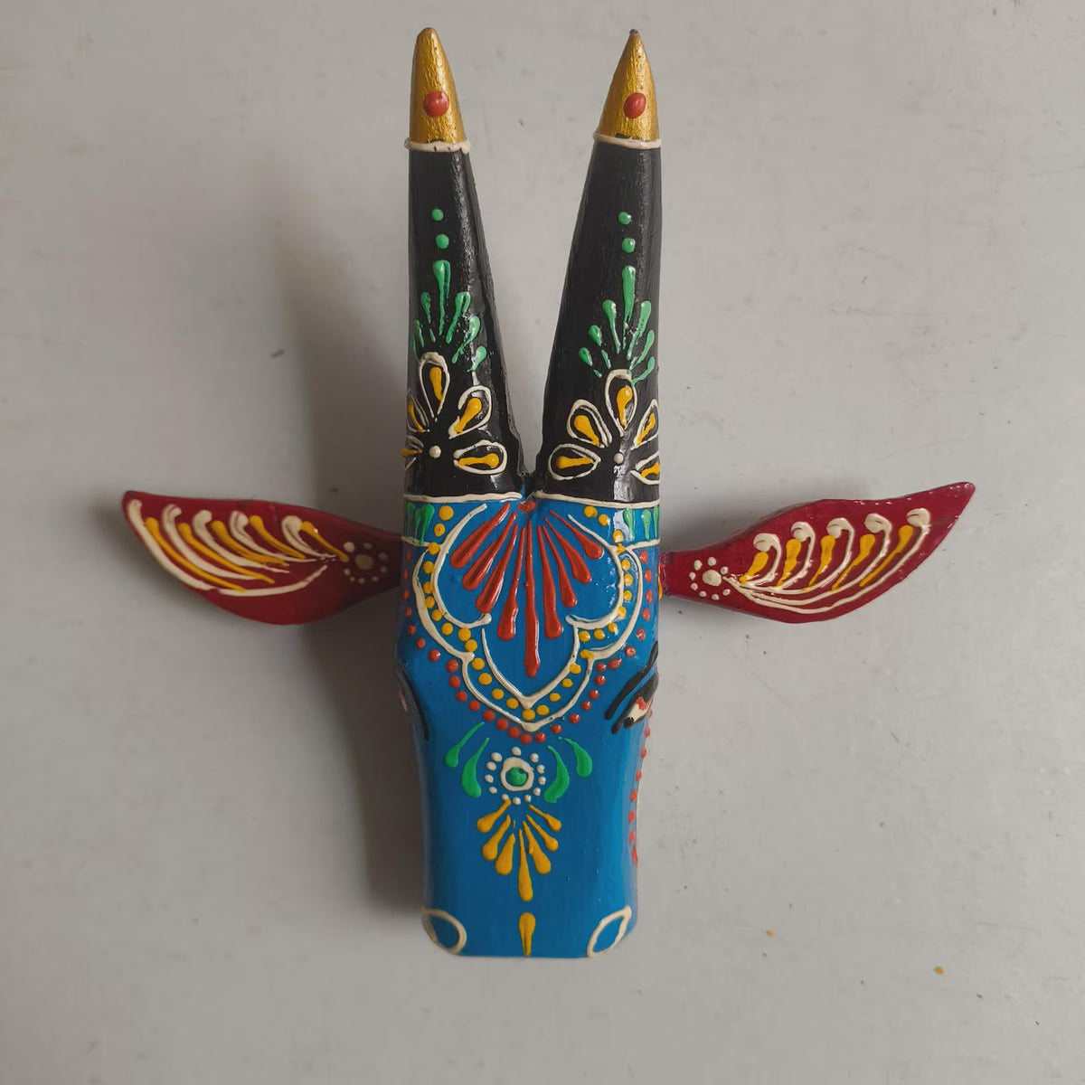A hand-painted wooden decorative object with two upright black horns tipped in gold, decorated with colorful floral and geometric patterns in blue, red, green, yellow, and white, with red wing-like side panels.