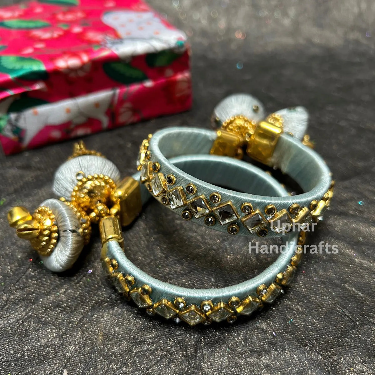 Grey silk thread bangles with Kundan stonework
Traditional bangle design with gold-tone embellishments
Decorative jhumka latkan hanging from the bangles
Metal base wrapped in colored silk thread
Indian-inspired jewelry for festive outfits
Part of a multicolor bangle collection
Uphar Handicrafts

