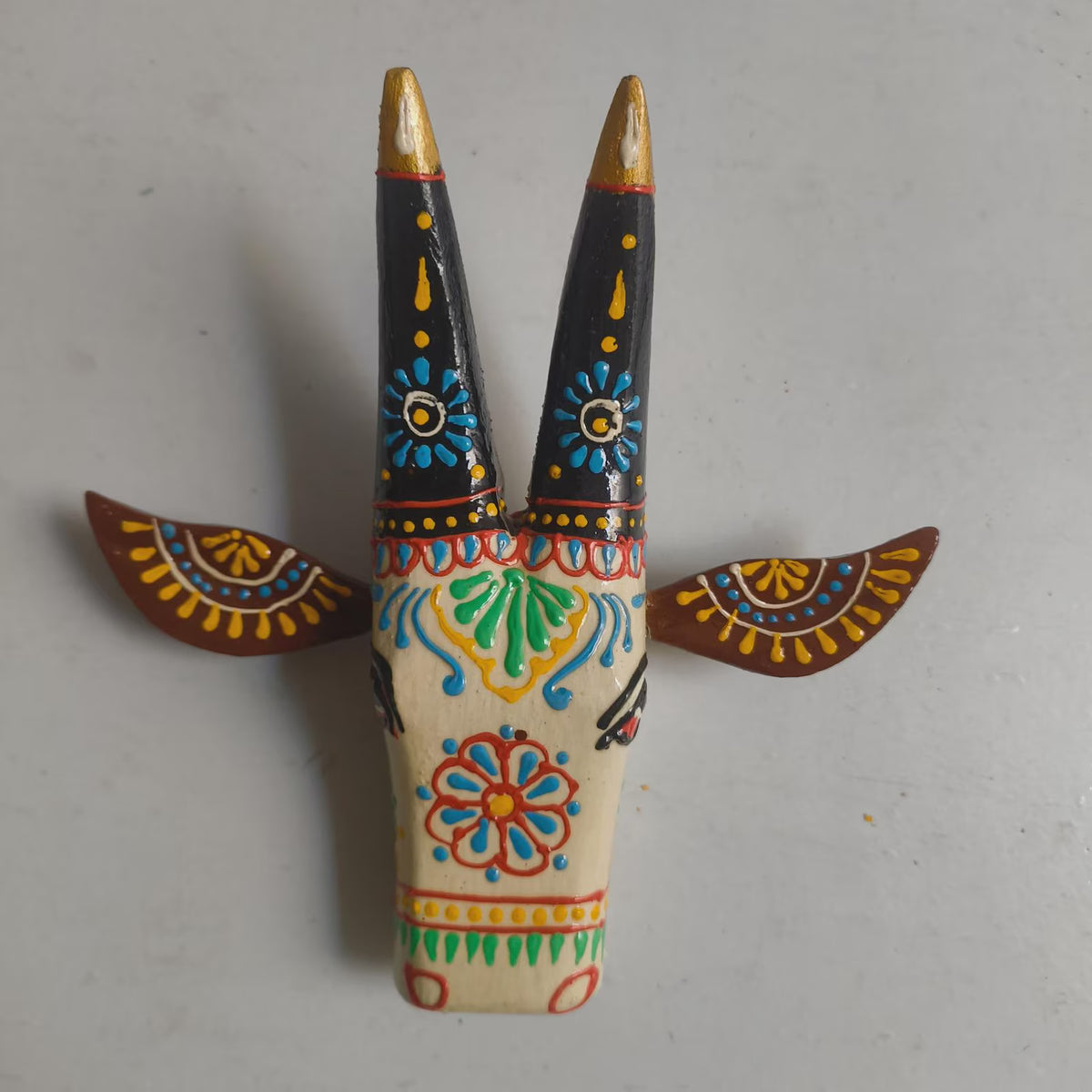 Hand-painted wooden Horn of God wall decor featuring a cream-colored face, black horns with gold tips, and colorful folk-art patterns on the horns and ears, set against a plain light gray background.