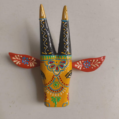 A hand-painted wooden folk art wall decor piece depicting a stylized goat head with tall black horns tipped in gold, a yellow face decorated with intricate multicolor patterns, and red ears with white and blue floral designs, set against a plain light gray background.