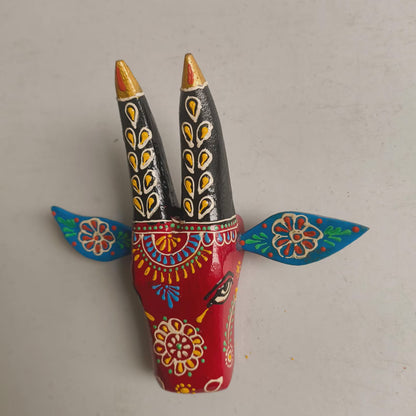 A hand-painted folk art figurine of a goat head, featuring a bright red body decorated with colorful floral patterns, black horns with gold and yellow details, and blue wings with red and yellow accents, set against a plain light gray background.